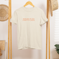 Beige t-shirt with 'meet me at the pumpkin patch' text on a wooden rack.