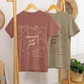 Two t-shirts, one brown and one green, hanging on a wooden rack with decorative items in the background.