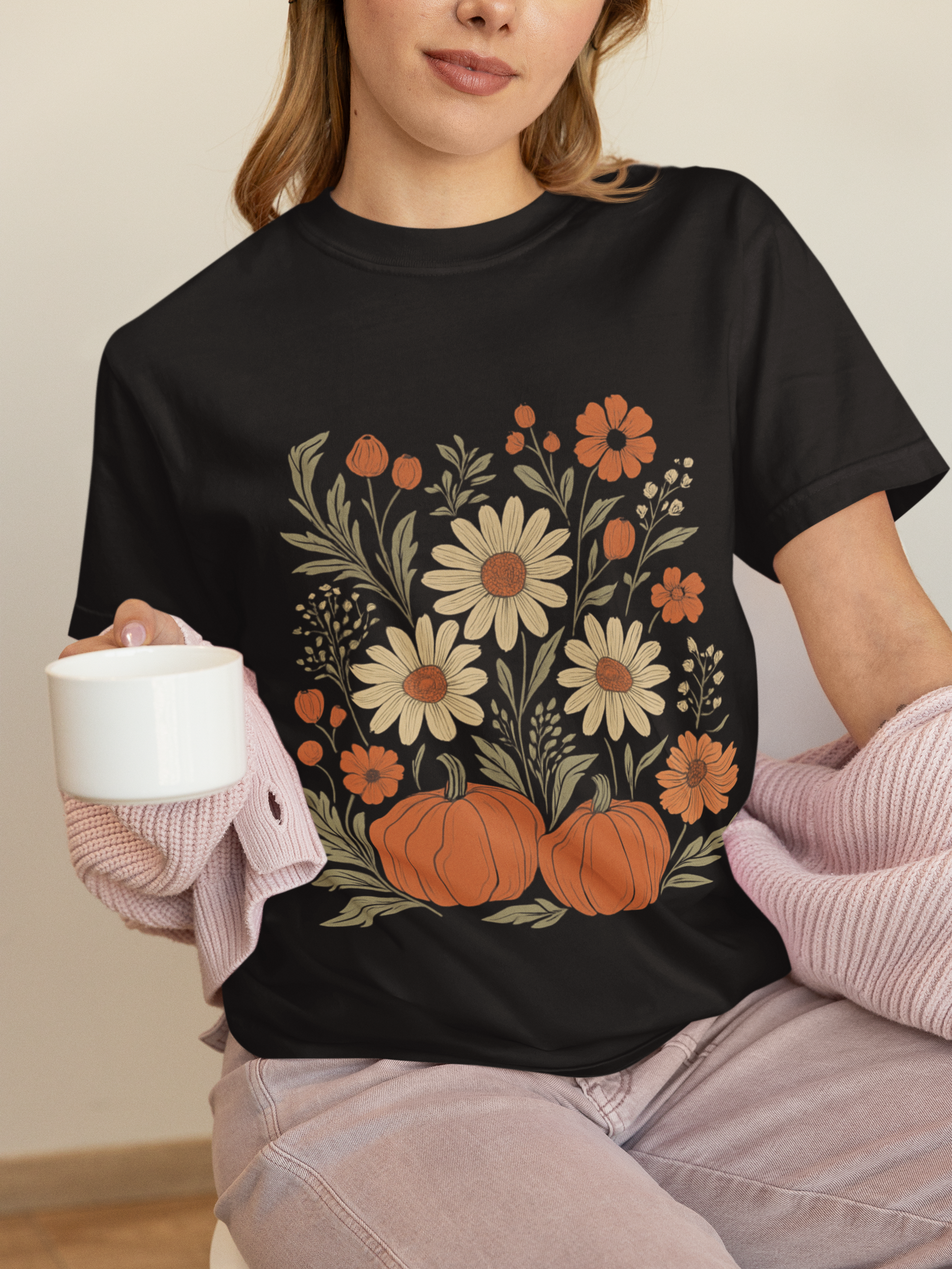 Person wearing a black t-shirt with floral and pumpkin design, holding a white mug.
